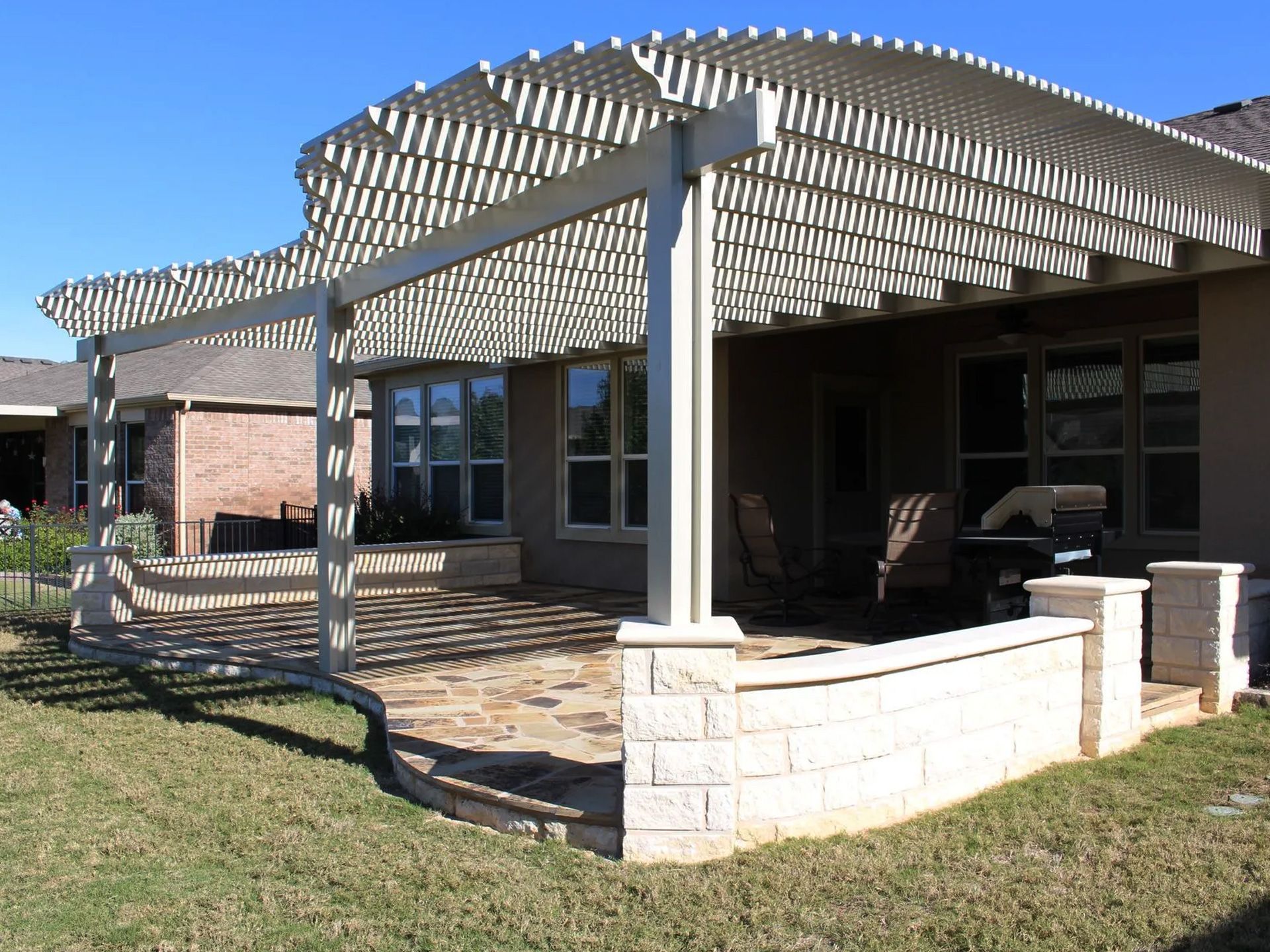 A pergola with horizontal slats covers a brick patio with a brick wall, overlooking a grassy yard.