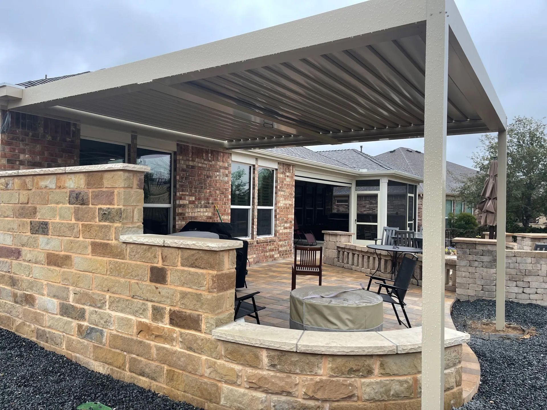 Patio with brick wall, tan pergola, seating, and fire pit in front of a house.
