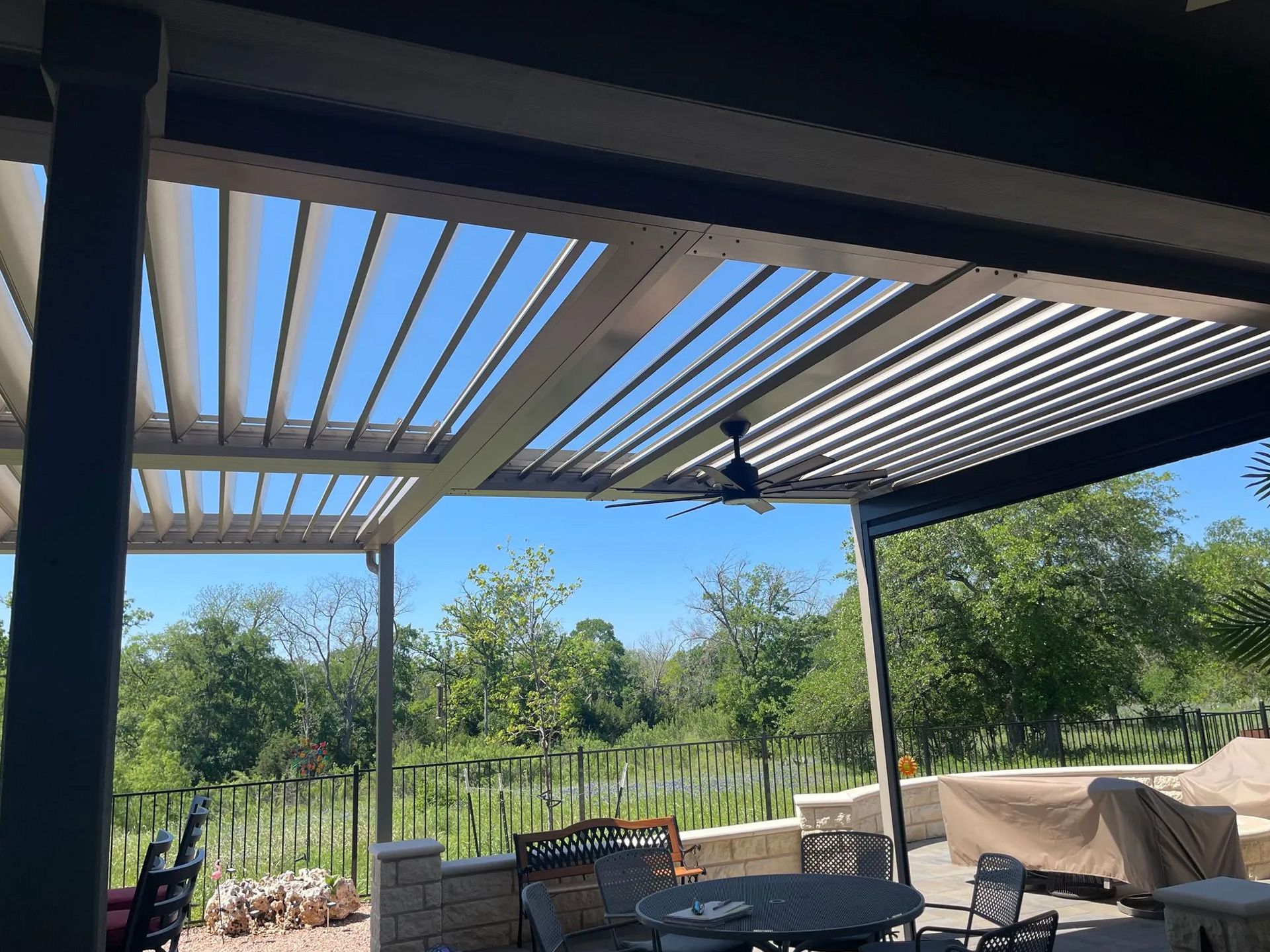 Patio with open-air pergola roof, outdoor furniture, and a ceiling fan. Sunny day with a view of trees.