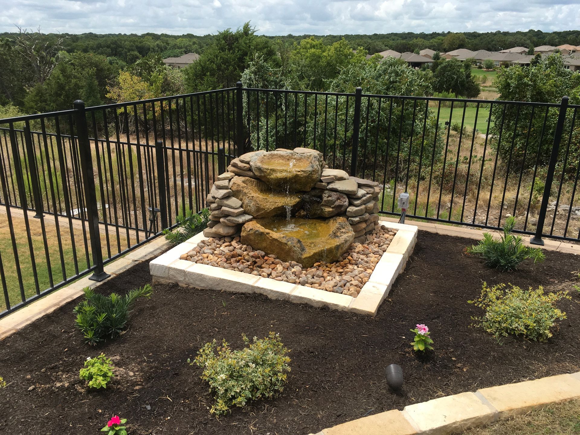 A water fountain is sitting in the middle of a garden next to a fence.