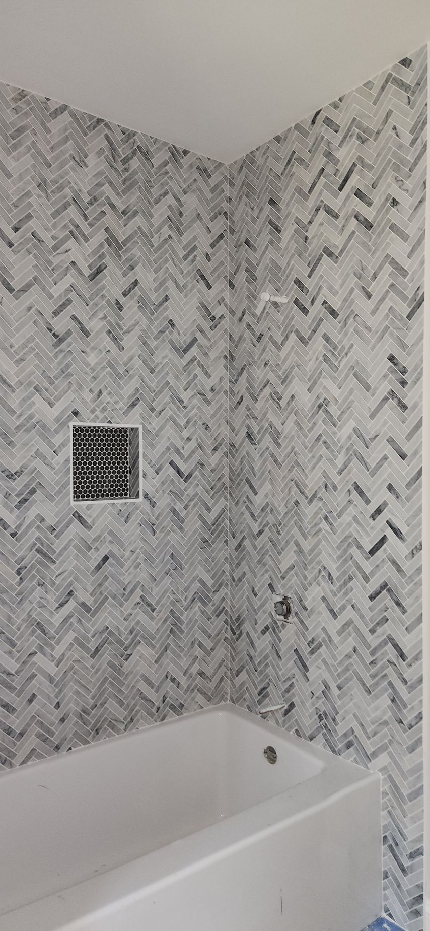 Bathroom with white bathtub, grey herringbone tile, and a small inset shelf.