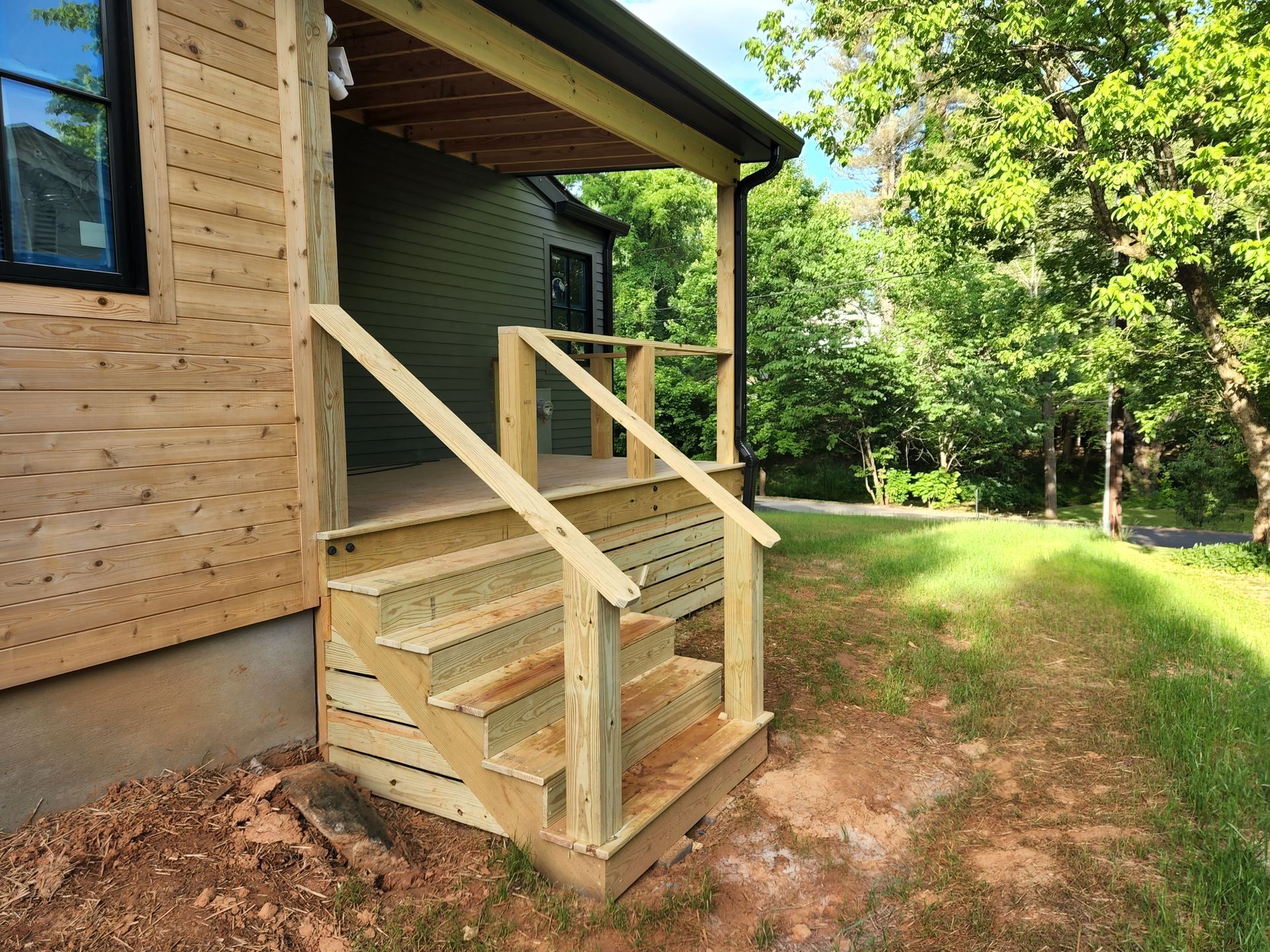 Wooden porch with steps and railing. Green house exterior with trees in background.