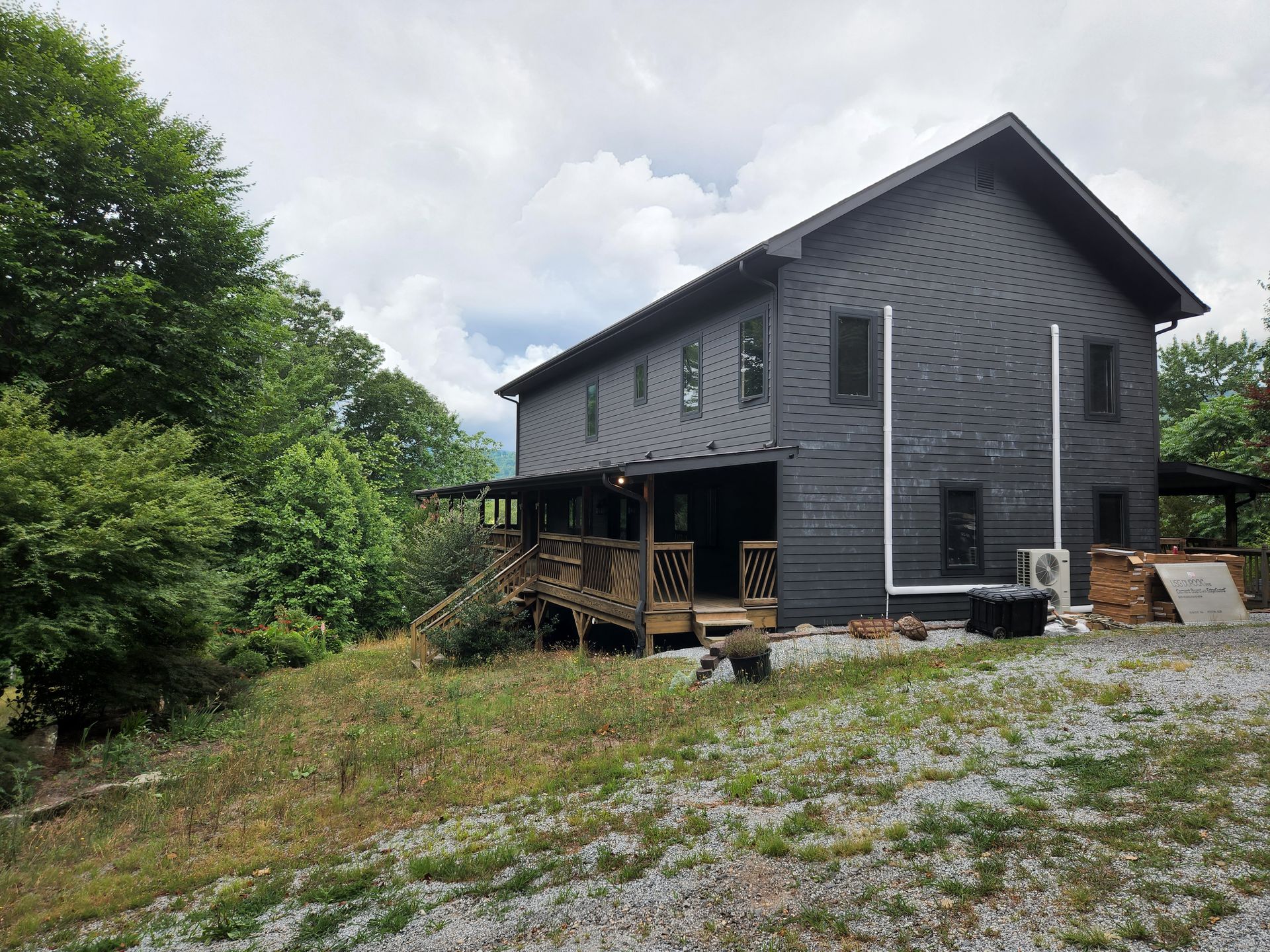 Dark gray two-story house with a porch, on a hillside, surrounded by trees, overcast sky.