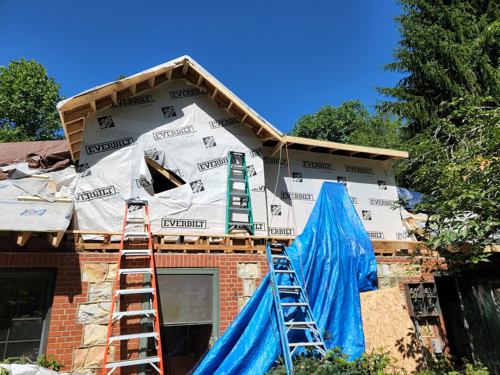 Construction in progress: brick house with new roof framing, covered with blue tarp and weather barrier. Ladders present.