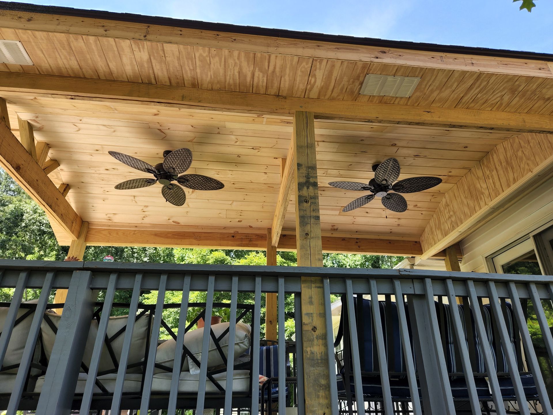 Deck with fans under a wooden ceiling, supported by posts. Gray railing and outdoor furniture visible.