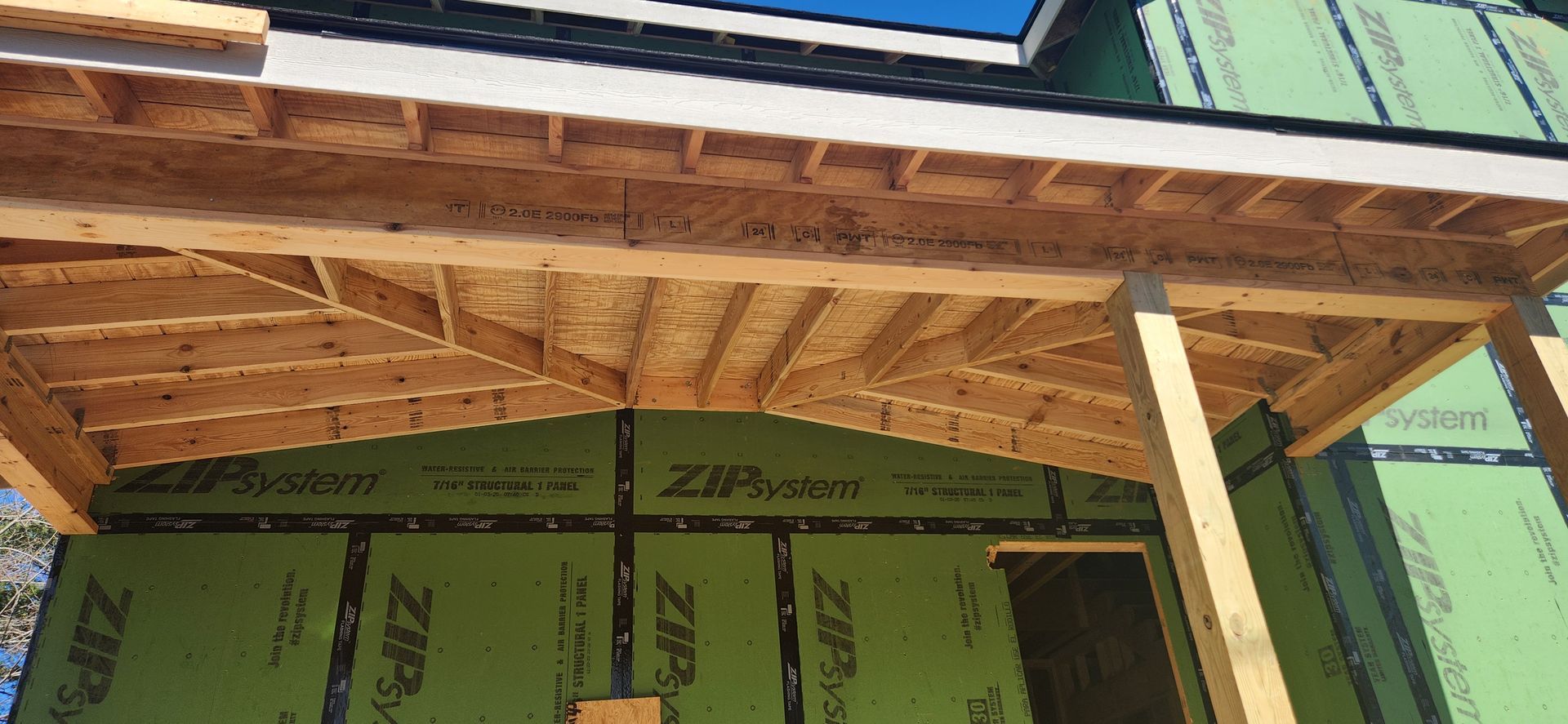 Wooden framing of a building's porch roof under construction. Green protective sheeting covers the walls.