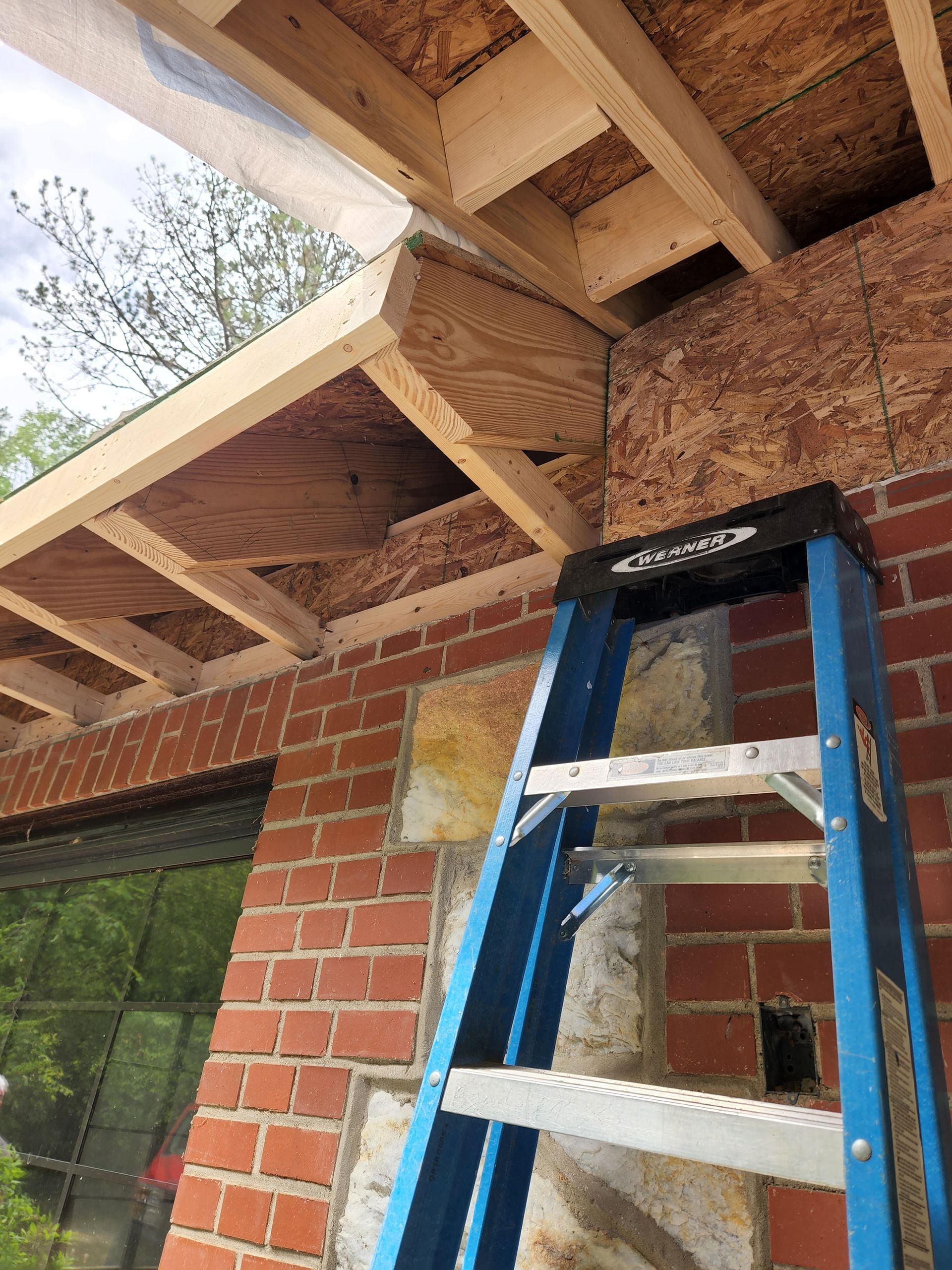Blue ladder propped against a brick wall; wood framing and OSB sheathing above, some covered with plastic.