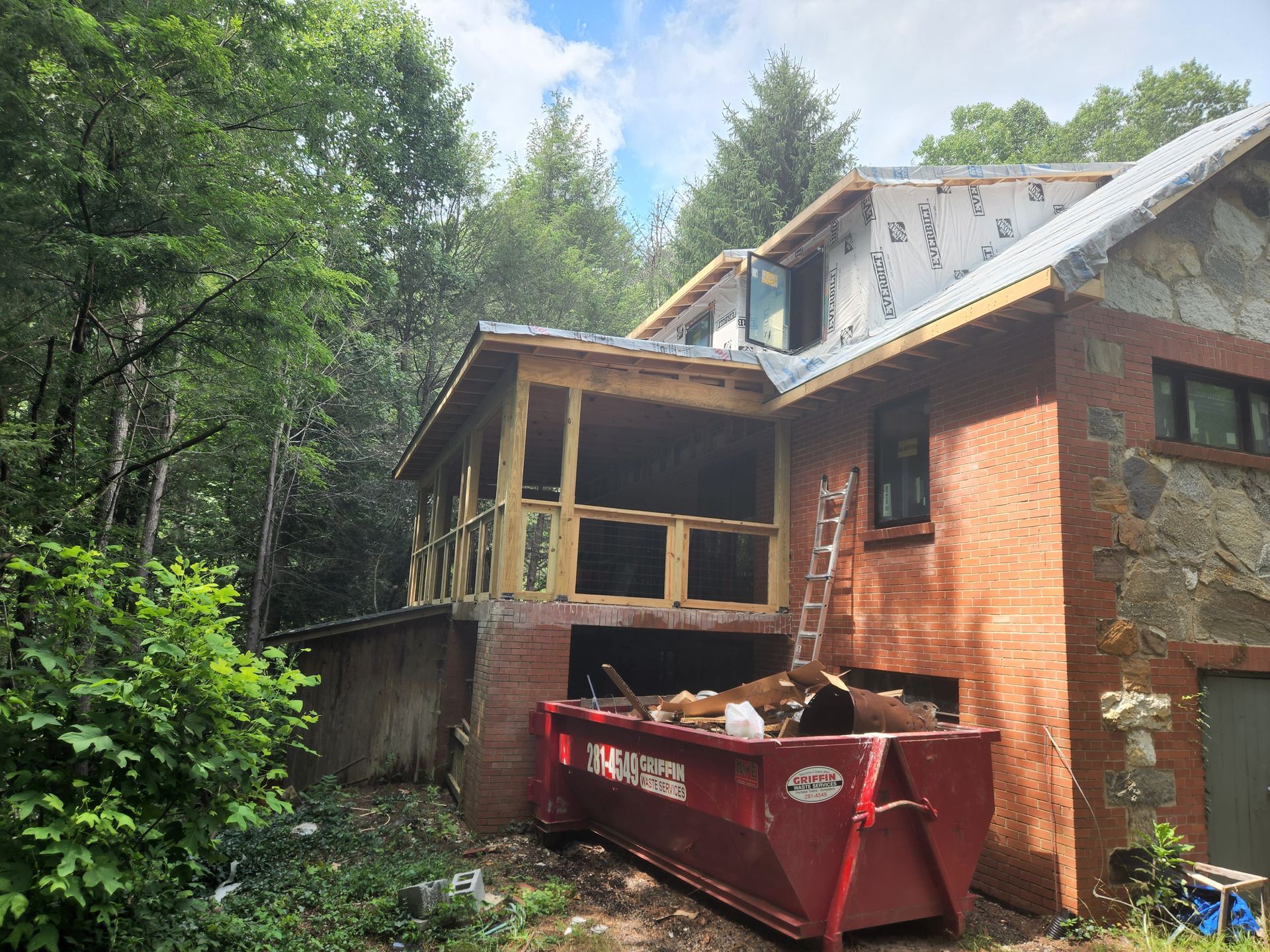 A two-story brick and stone house under construction, with a red dumpster in front.
