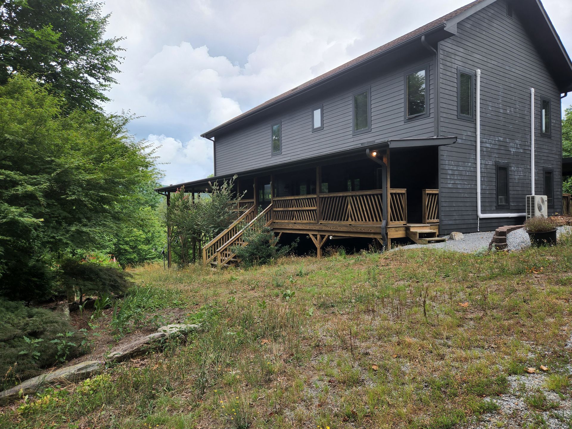 Two-story dark house with wooden porch and deck on a grassy hill, surrounded by trees.