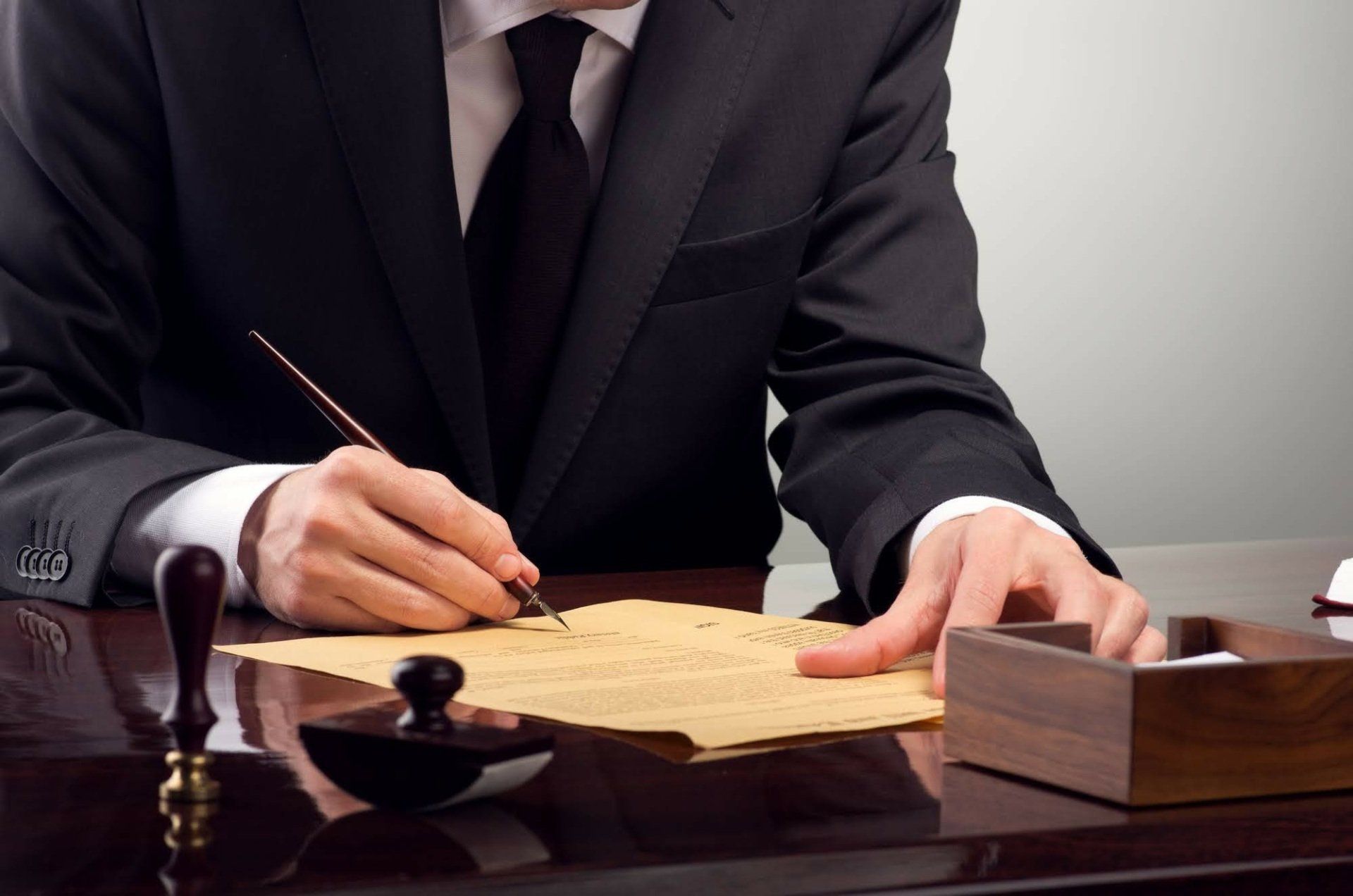 Man in a suit writing on aged paper with a quill pen at a desk. Seal and wooden box present.