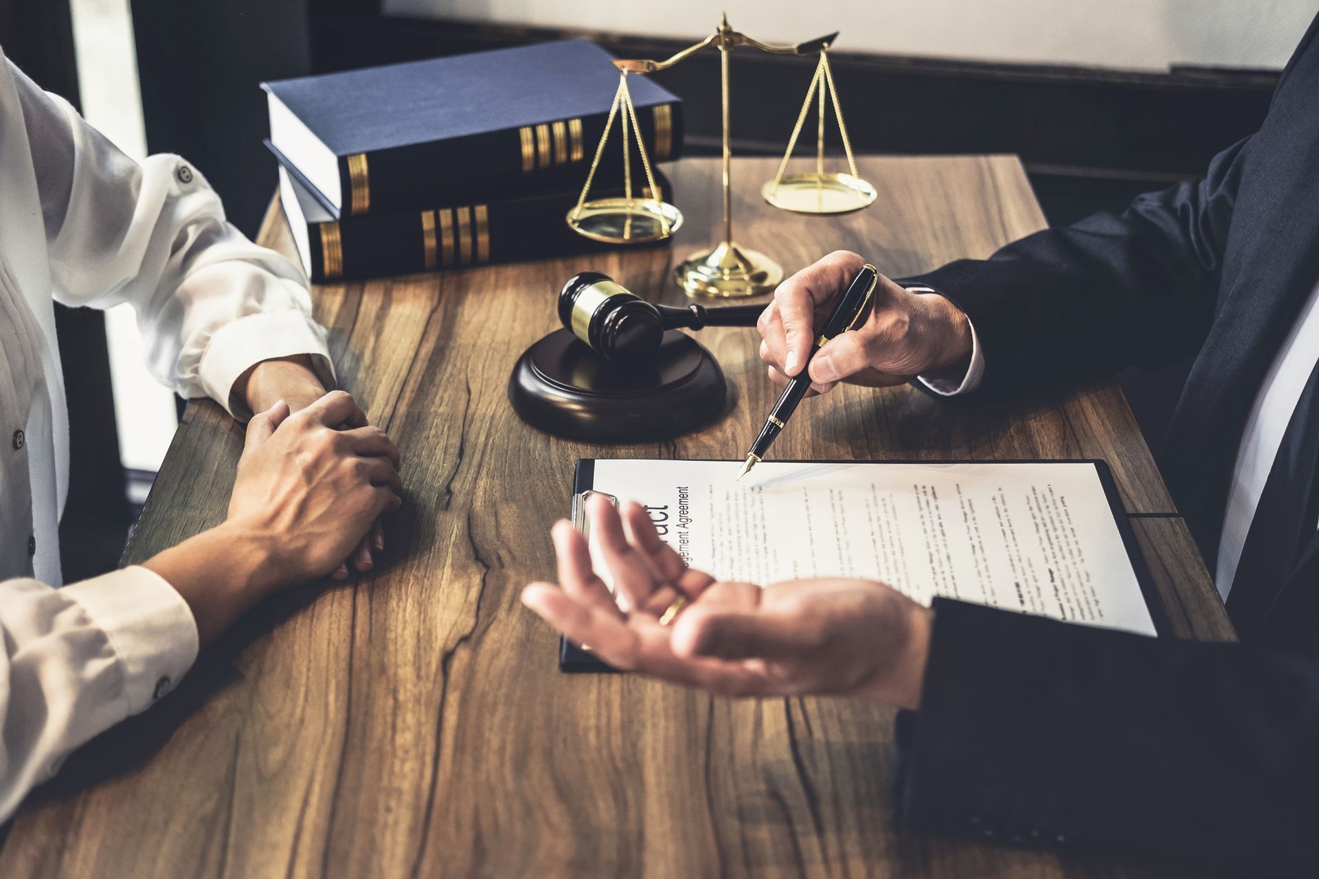 Lawyer in suit pointing to document, consulting with a person. Law books, gavel, and scales of justice on the table.