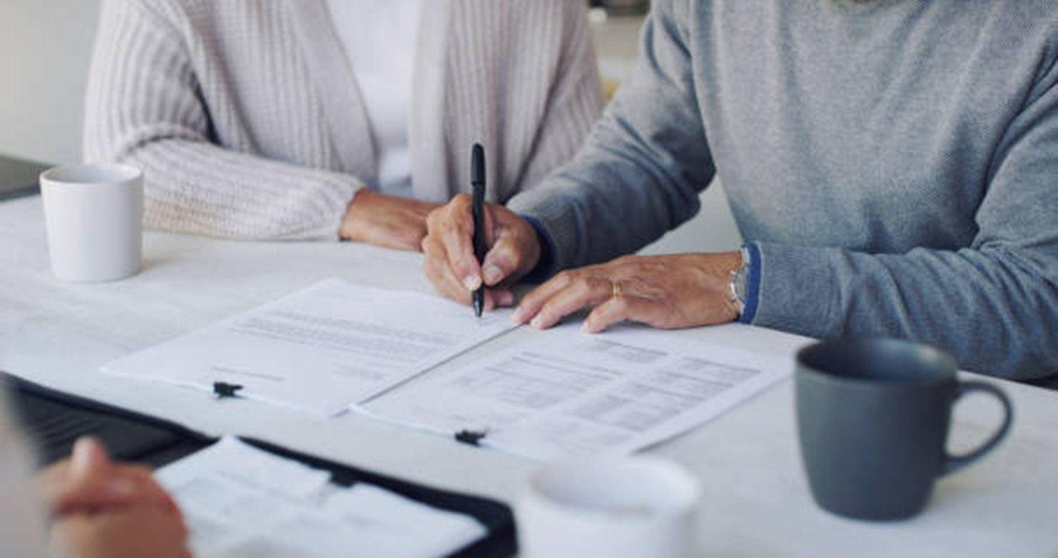 Person signing documents at a table; another person watches. Cups and papers are visible.