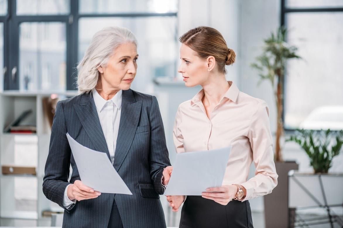 Two women in an office looking at papers, talking. One has gray hair, a suit, and the other is younger, wearing a button-down shirt.