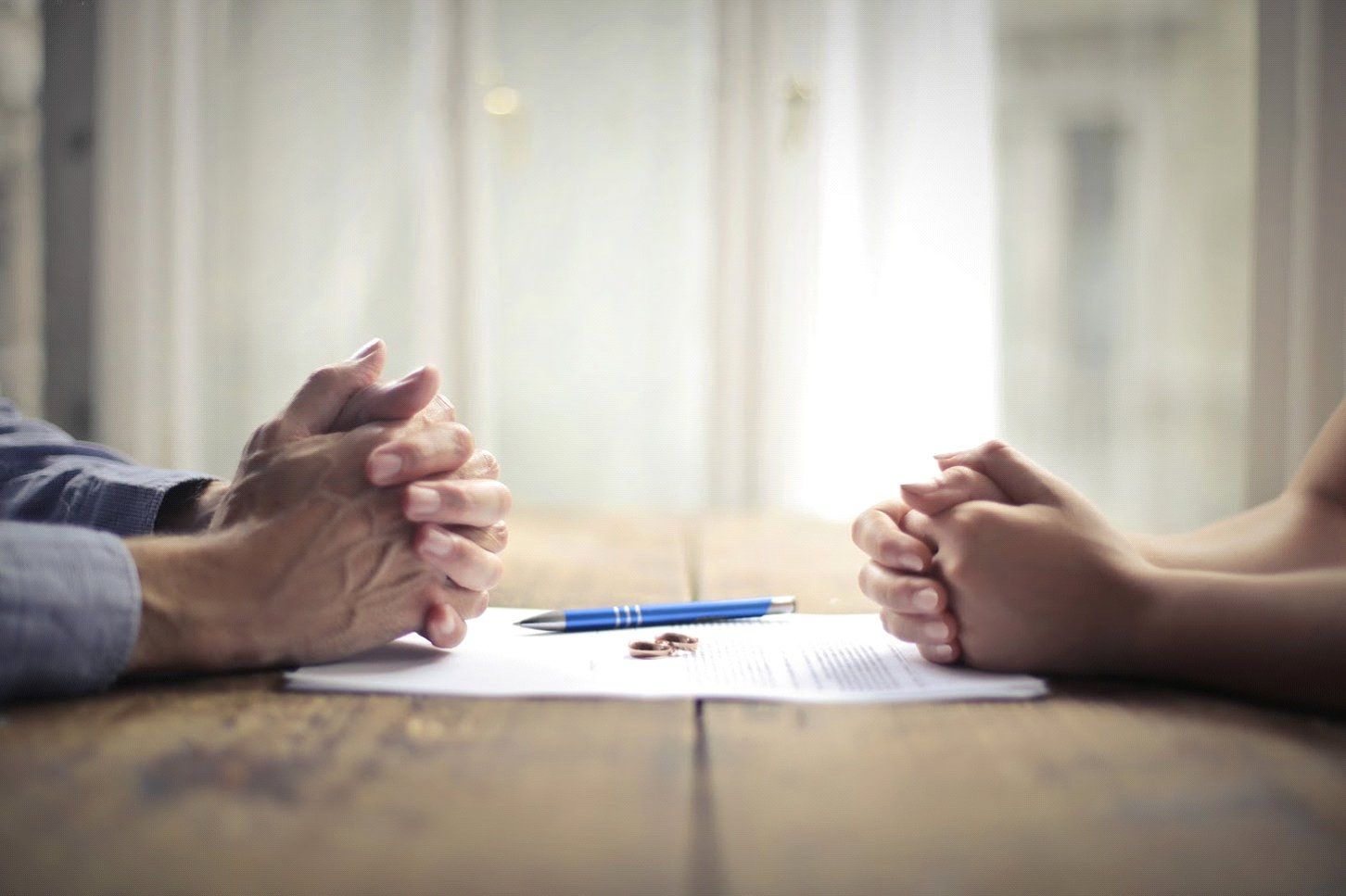 Hands clasped on a table with a pen, divorce papers, and a wedding ring.