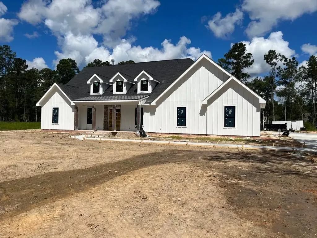 New white farmhouse under construction with black roof, blue sky, and trees.