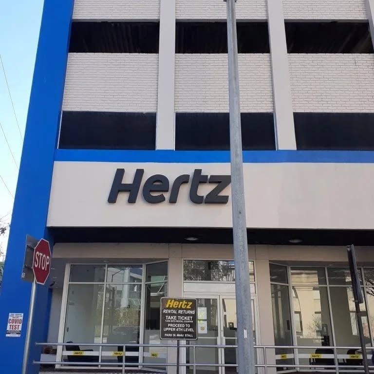 Hertz rental car building with large black sign. Blue and white exterior. Stop sign in front.
