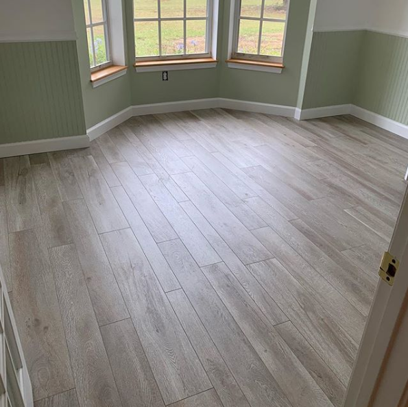 Empty room with light wood-look flooring, pale green walls with wainscoting, and a window seat with windows.