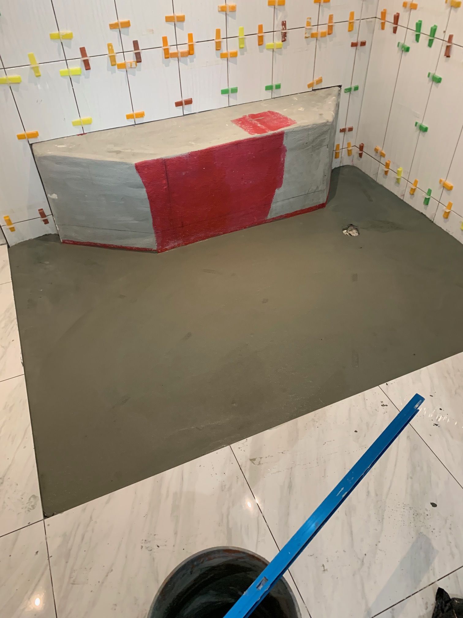 Bathroom under construction, gray cement floor, tiled walls, large red-covered form, blue tool, bucket with dark liquid.