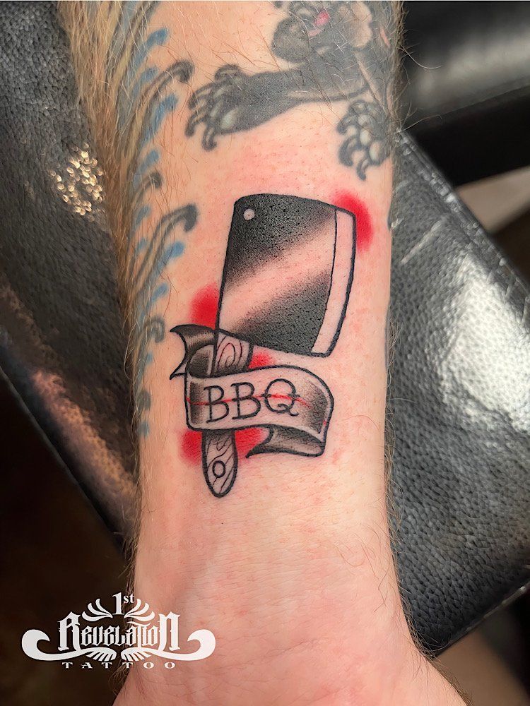 A tattoo of a cleaver with a banner that says bbq on it on a person's wrist.