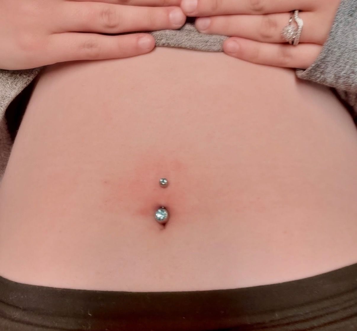 Close-up of a person's abdomen with a double belly button piercing, hands above.