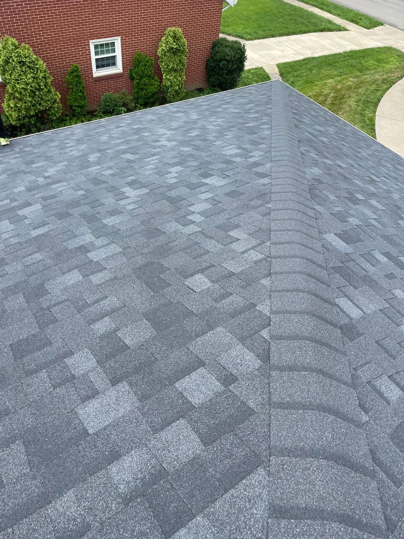 Gray asphalt shingle roof with signs of weathering, view from above. Background includes a brick building and green lawn.