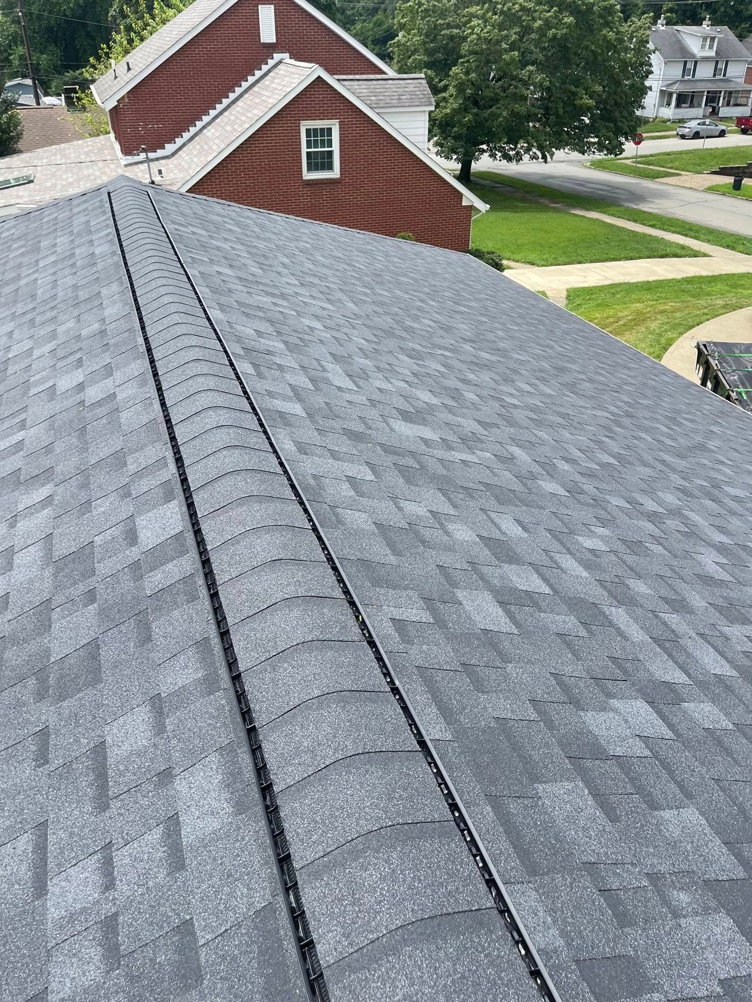 View from a roof, featuring gray asphalt shingles and a peak. A red brick building is in the background, along with green grass and houses.
