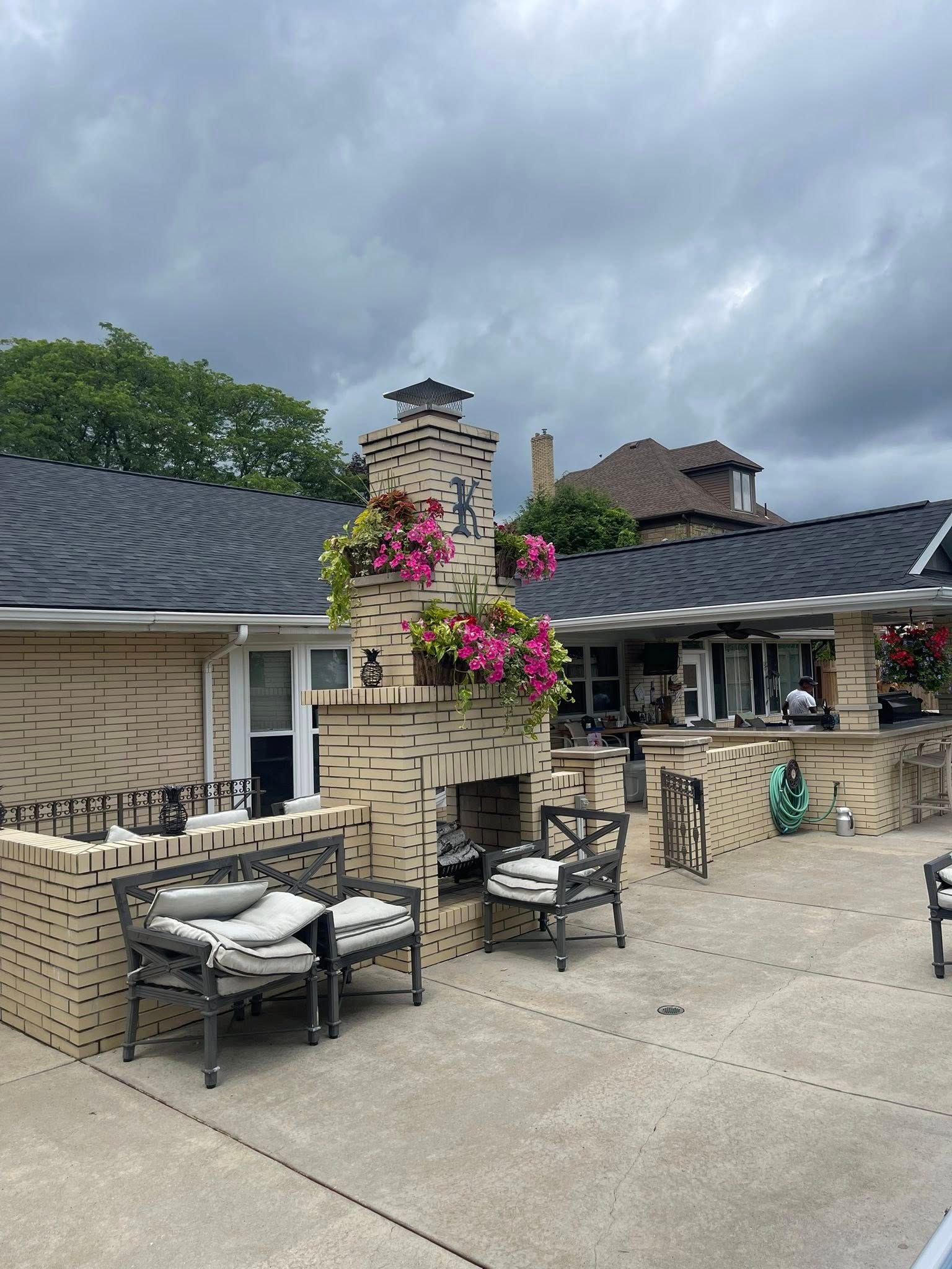 Outdoor patio with brick fireplace adorned with hanging pink flowers, seating, and a gray sky.