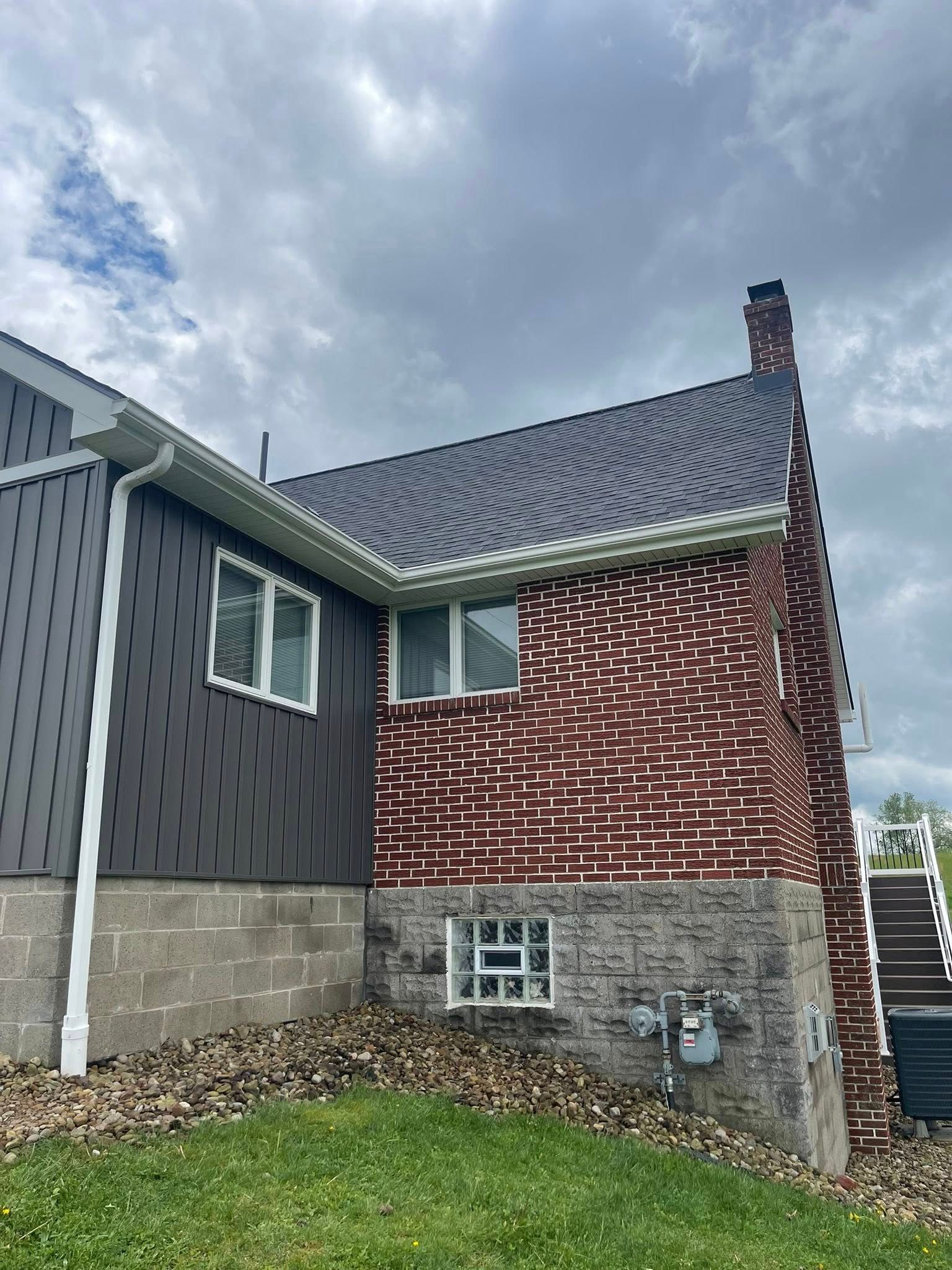 Side view of a house with dark gray siding and a brick chimney. The roof is dark, and the overcast sky is visible.