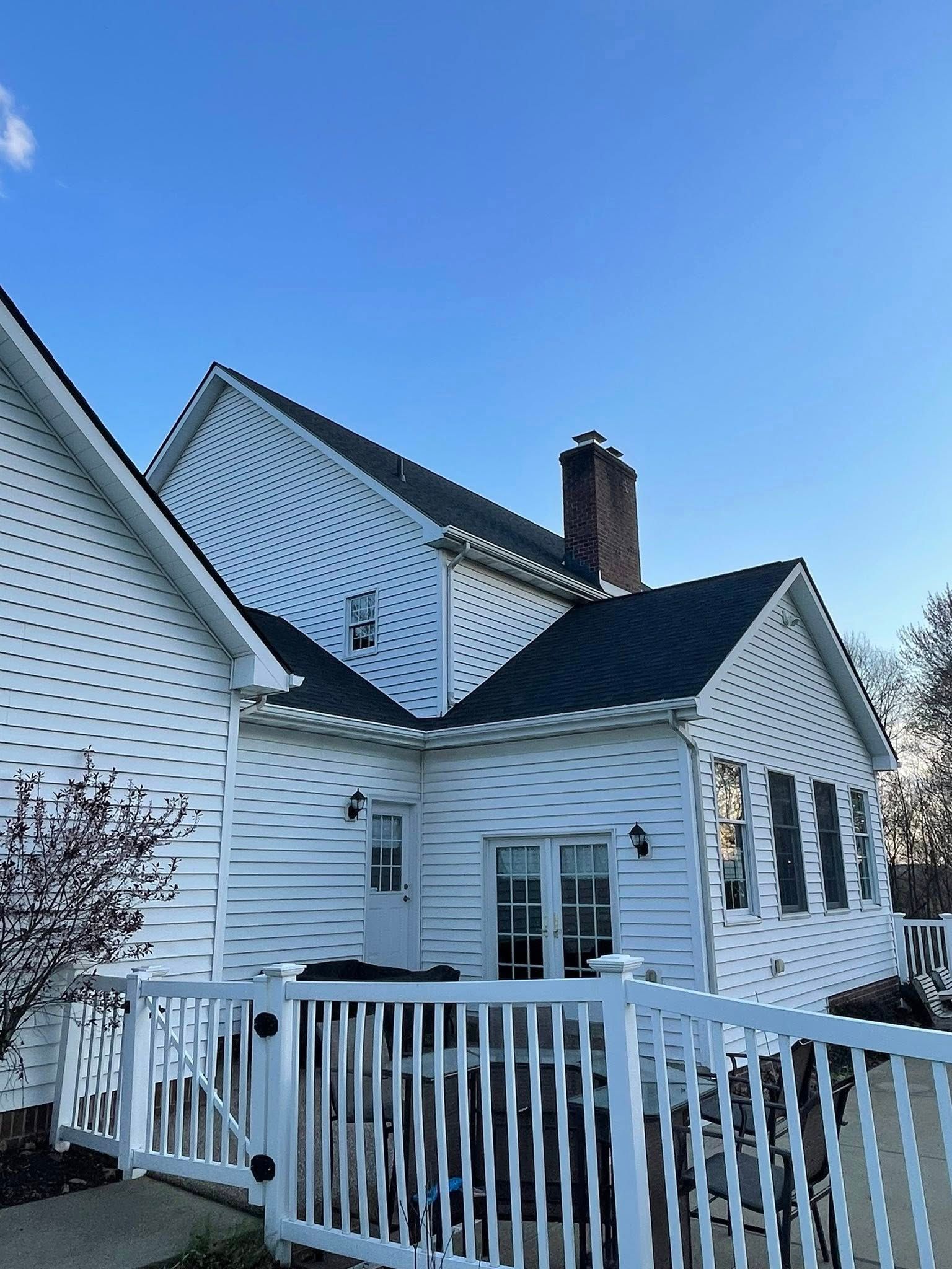 White house with black roof and chimney against a blue sky. A white fence and small patio are in the foreground.