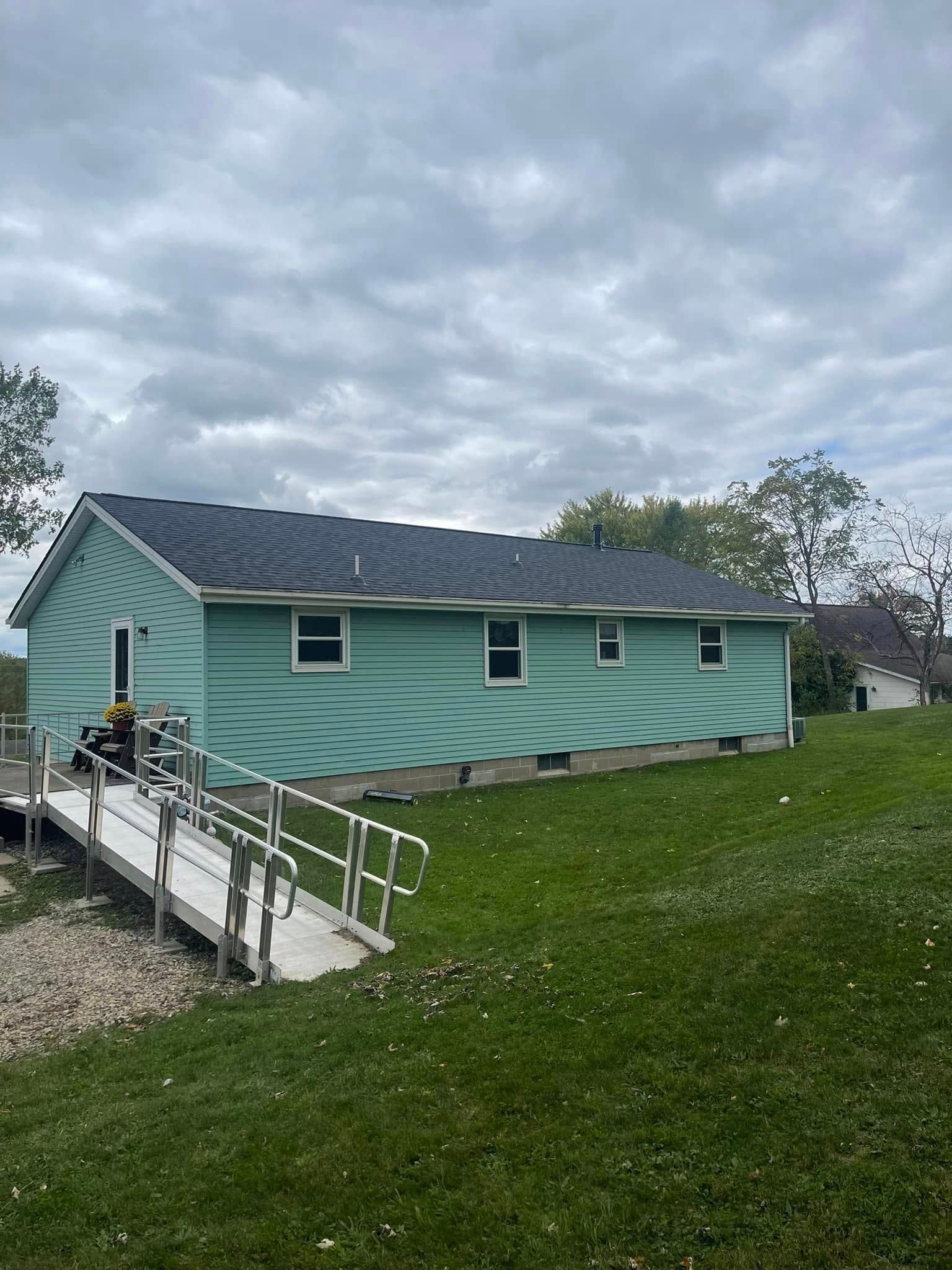 Exterior of a light green building with a dark roof, accessible ramp in foreground, on a grassy hill under a cloudy sky.
