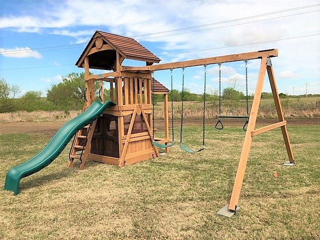 Wooden backyard playground with green slide and swings on a grassy field under a blue sky