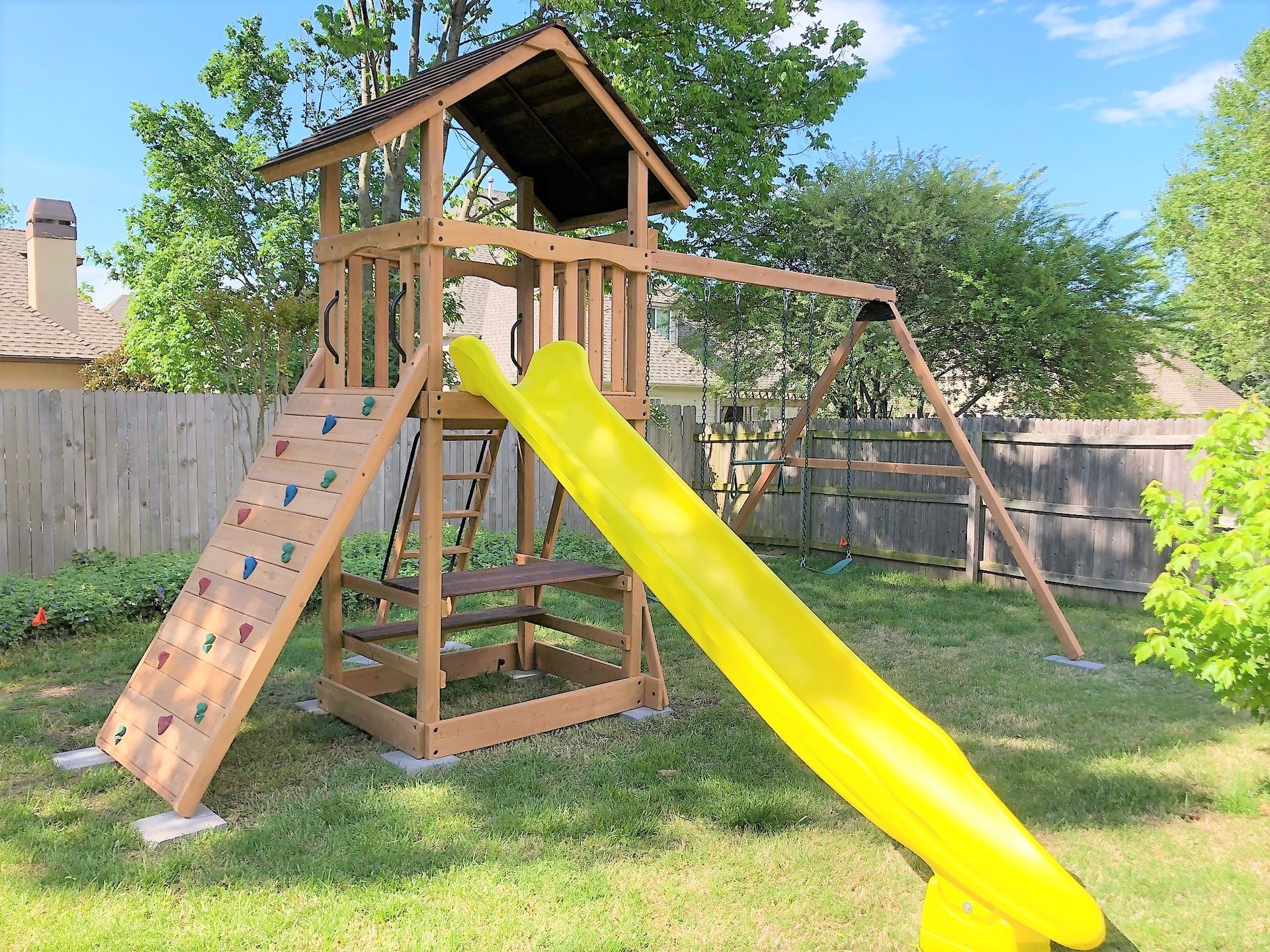 Backyard wooden playset with yellow slide, climbing wall, and swing under a blue sky