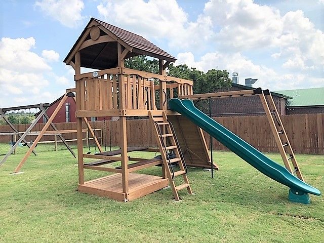 Wooden playground with a slide, swing set, and roofed play deck on a grassy field.