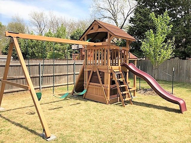 Wooden backyard playground set with swings, slide, and climbing deck in a fenced yard