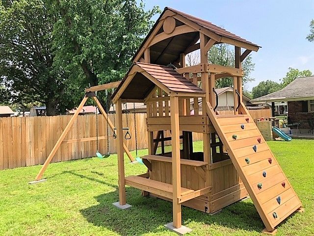 Wooden backyard playset with climbing wall, roofed platforms, and slide in a fenced yard