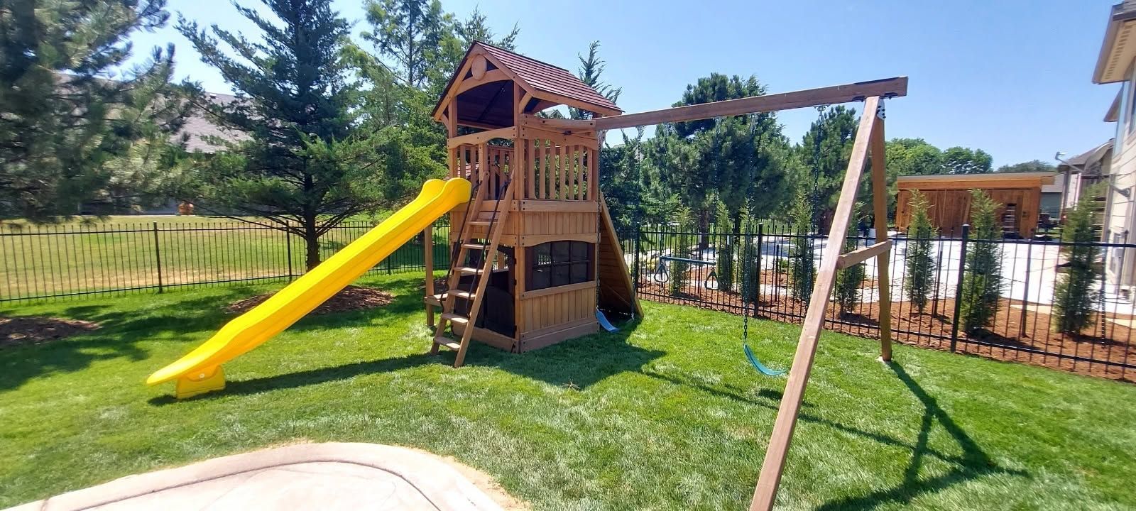 Wooden playground playset with a yellow slide and swings on a grassy lawn under a blue sky