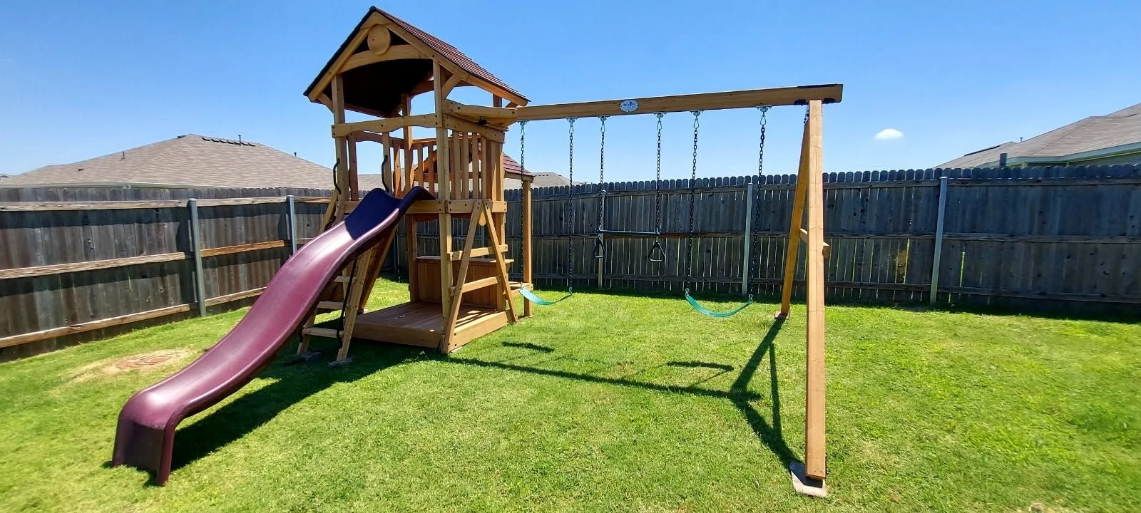 Backyard playset with purple slide, wooden tower, and swing set on a grassy lawn.