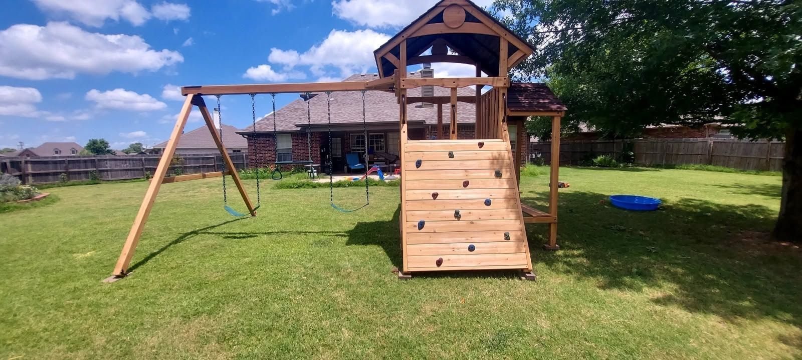 Backyard wooden playset with swings and climbing wall on a grassy lawn under a blue sky