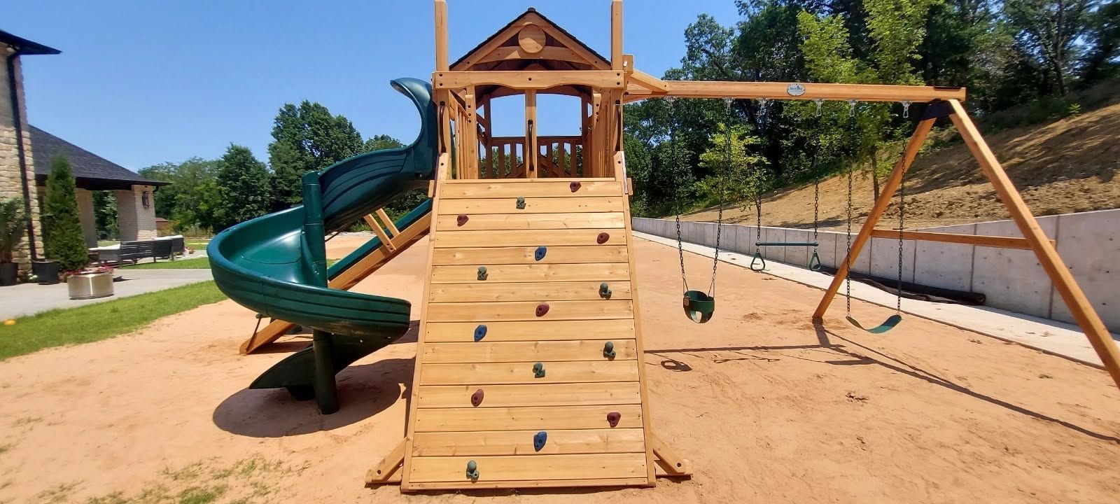 Children’s playground with wooden climbing wall, green slide, and swings on sand outdoors