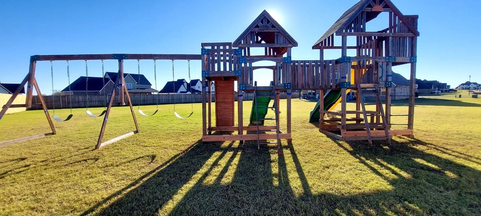 Wooden playground structures and swings on a grassy field under a bright blue sky.