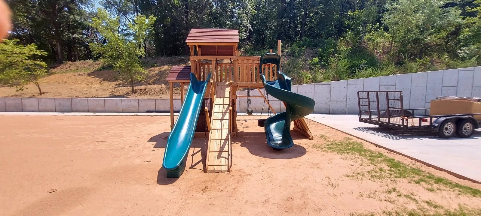 Playground with a blue slide, climbing structure, and spiral slide on a sandy lot beside a trailer.