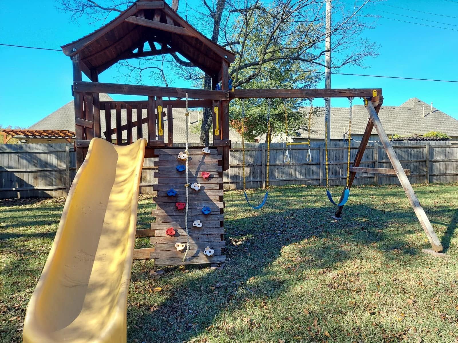 Backyard playground with yellow slide, climbing wall, swing set, and wooden playhouse in a fenced yard