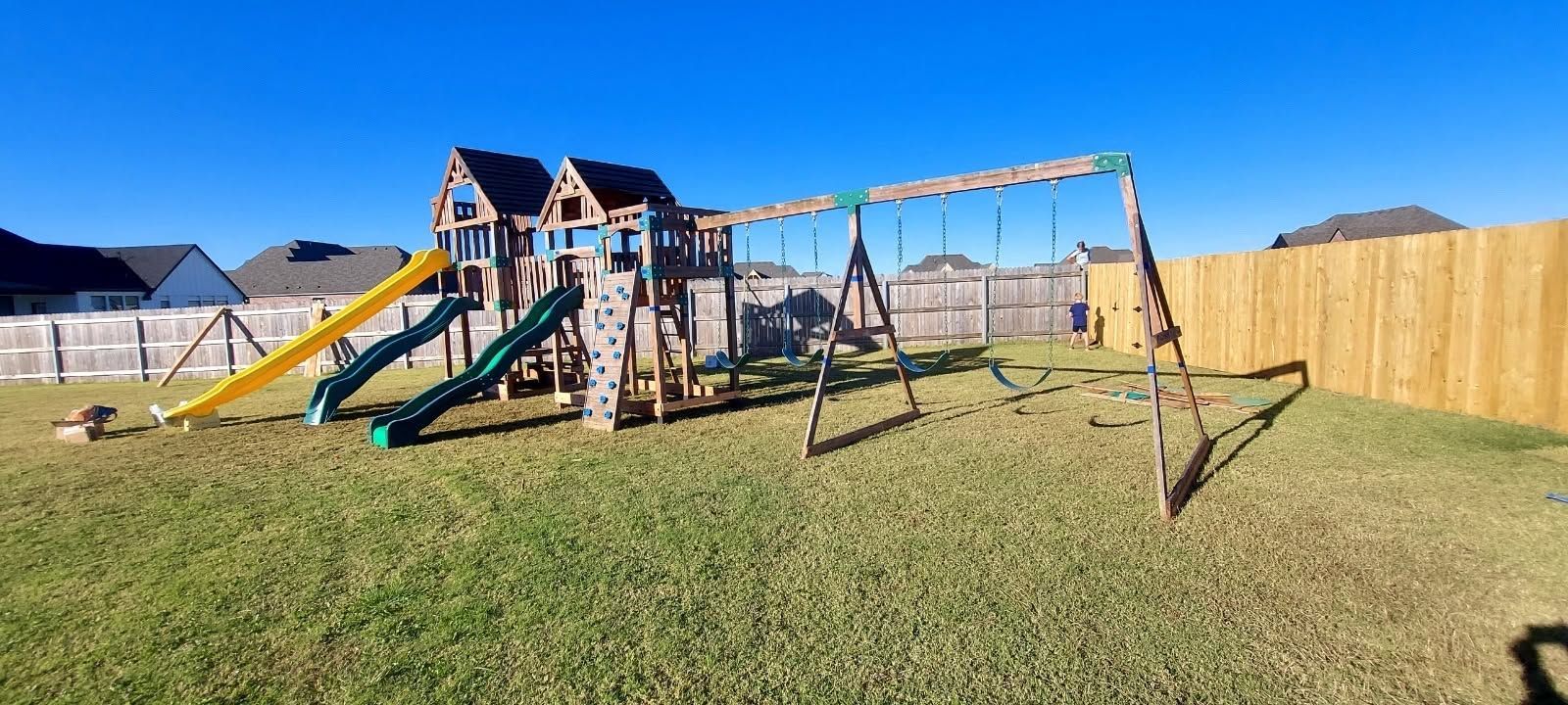 Backyard playset with slide, swings, and climbing frame on a grassy lawn under a clear blue sky