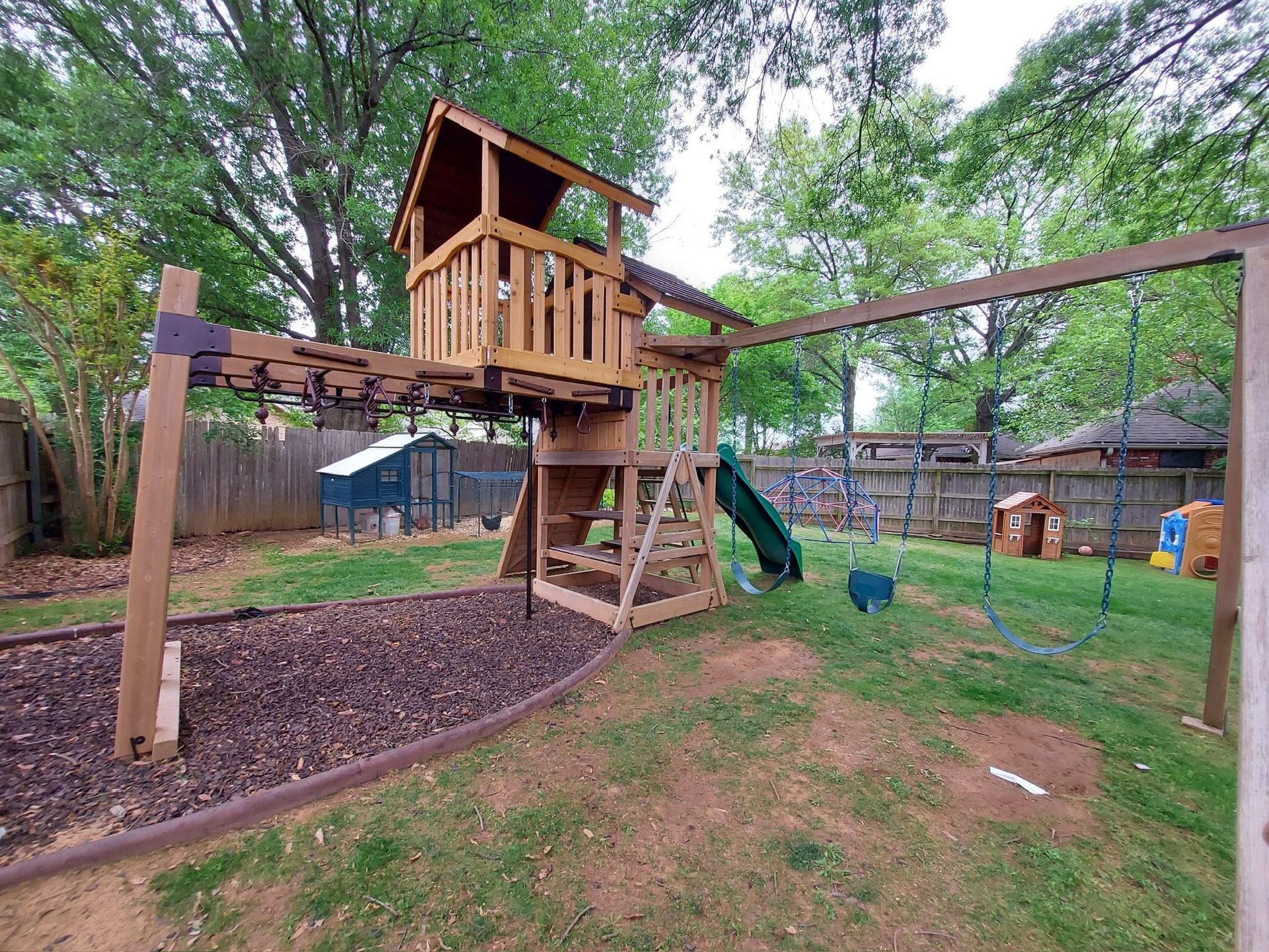 Wooden backyard playset with treehouse, swings, and slide under green trees