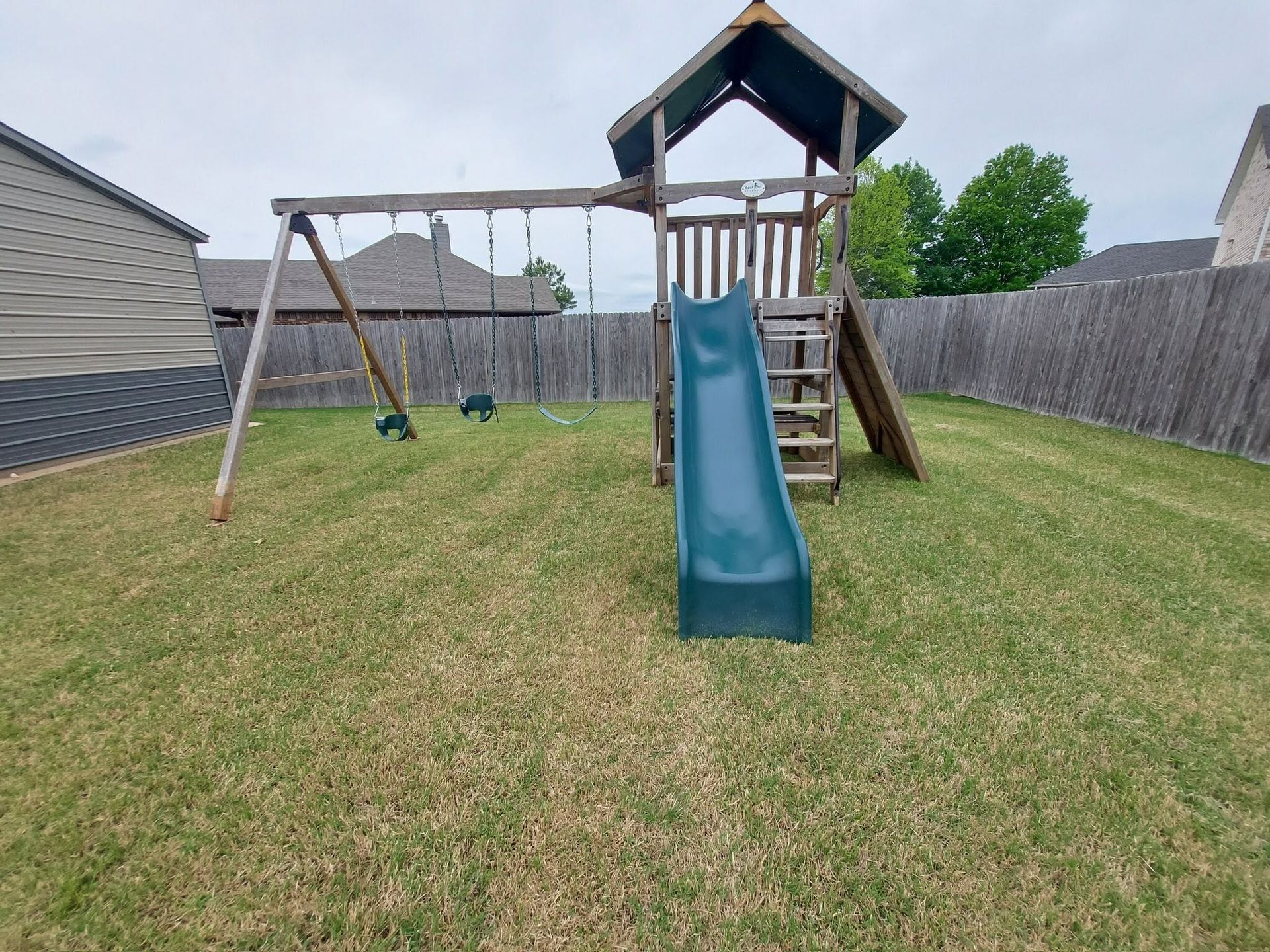 Backyard playset with blue slide, swing set, and ladder on a grassy lawn between fenced houses