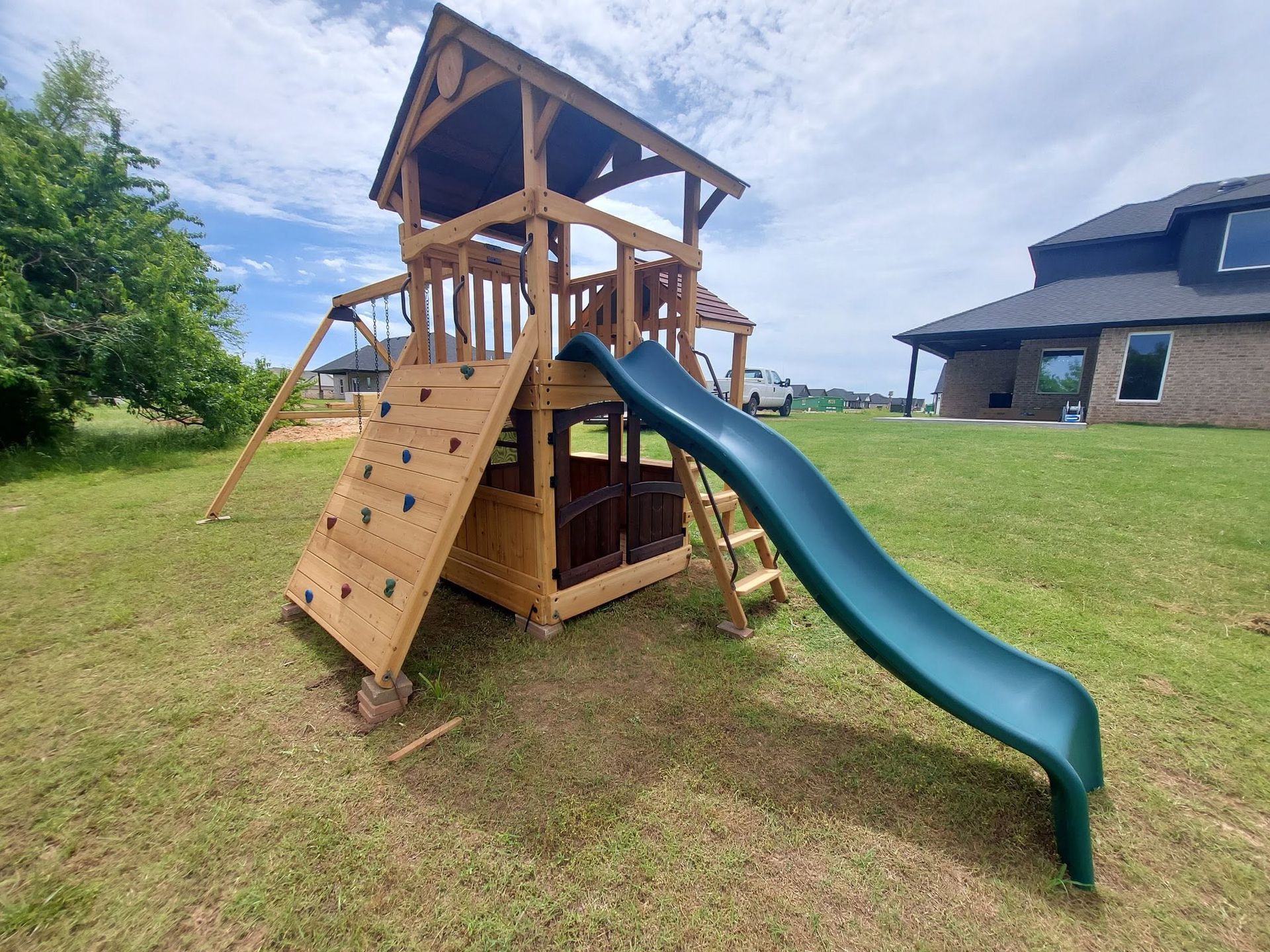 Wooden backyard playset with climbing wall, slide, and roofed tower on a grassy lawn.