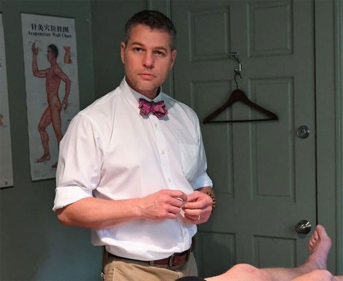 Man in white shirt and bow tie stands in a medical exam room, adjusting his hands.