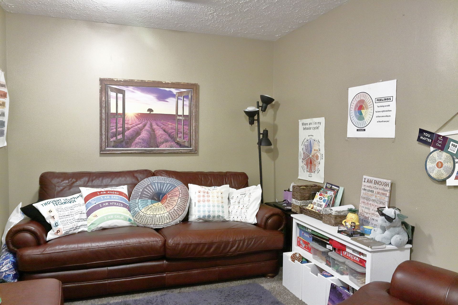 A living room with a brown couch and a painting on the wall. - Beth