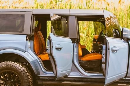Light blue off-road SUV with open doors and orange seats parked in tall grass