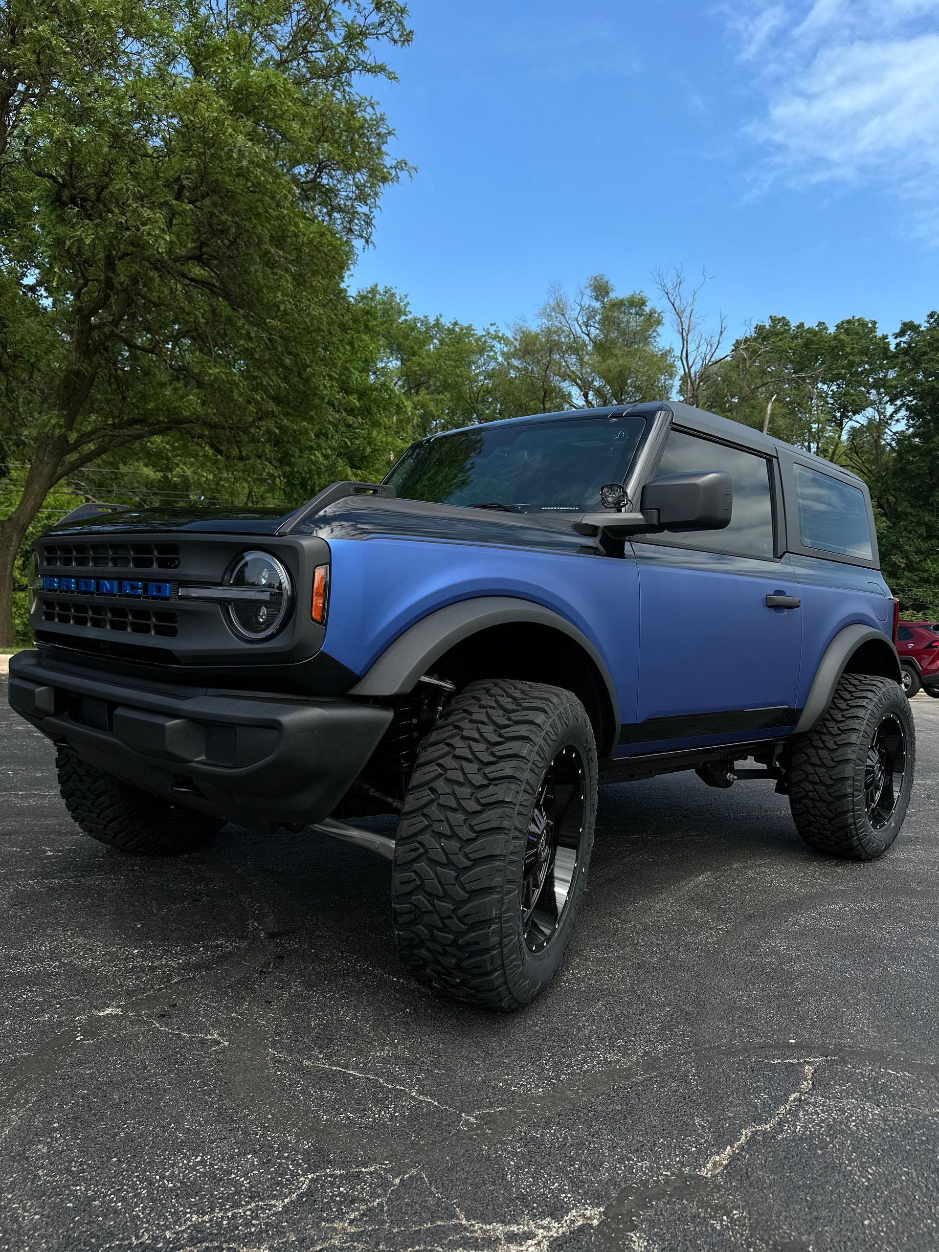 Blue off-road SUV with large tires parked on gravel near trees under a blue sky
